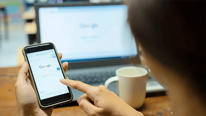 Close up of a mobile phone on Google site held by a hand and a laptop on the background