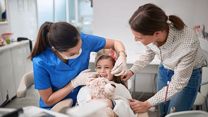 A young child sits in the dental chair, receiving care from a dentist, while the mother observes the procedure.