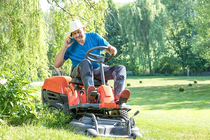 Man with a grass trimmer mows the lawn while holding a mobile phone to his ear