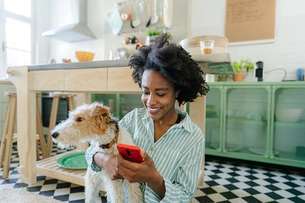 A girl sitting on the kitchen floor, holding a dog as she uses her phone