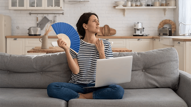 Woman sitting on her couch feeling warm inside her home, holding a fan to keep her cool