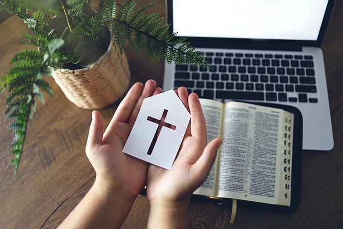 Image of hand holding a house-shaped paper with a cross cutout with a Bible and a laptop in the background. 