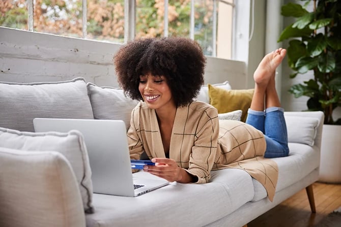 Smiling young African American woman lying on her living room sofa scheduling a salon appointment for hair color.