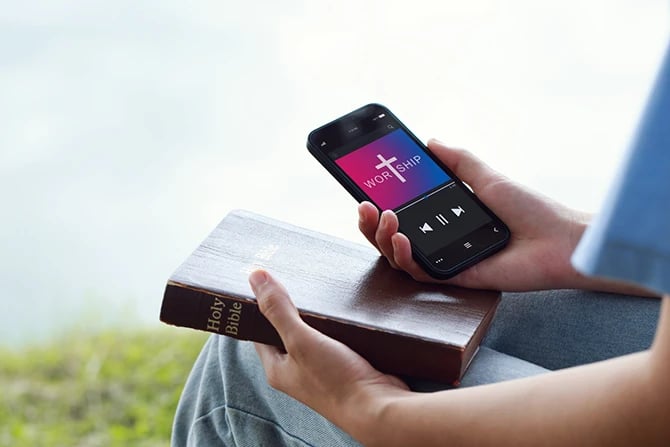 Close-up of a young man’s hands holding a Bible in the left hand and his mobile phone livestreaming a worship service.