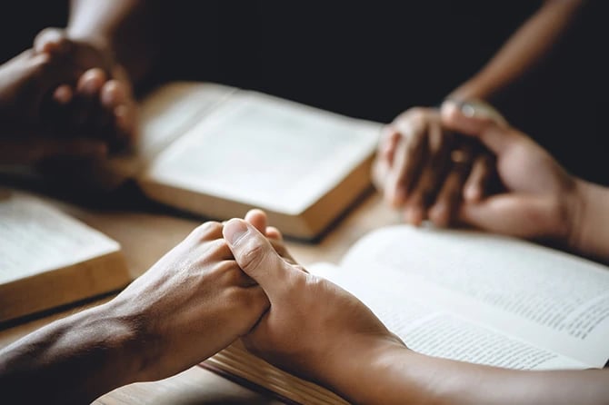 Close-up shot of several hands holding each other while they have their bibles open in front of them.