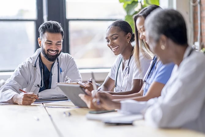 A group of four medical professionals dressed in scrubs and lab coats meeting around a boardroom table.